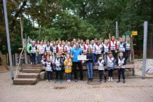 Eine große Gruppe Grundschulkinder in Warnwesten steht mit zwei Erwachsenen auf einem Schulhof, eine Frau hält eine Urkunde.