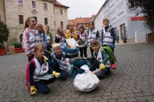 Eine Gruppe Grundschulkinder in Warnwesten sitzt und steht mit gefüllten Beuteln und Greifzangen auf einem Kopfsteinpflasterplatz vor historischen Gebäuden.