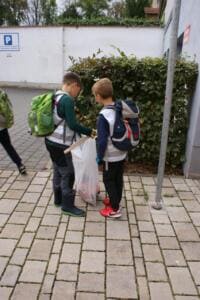 Zwei Jungen mit Schulranzen und Warnwesten sammeln mit Greifzange und Beutel Müll an einer Hecke.