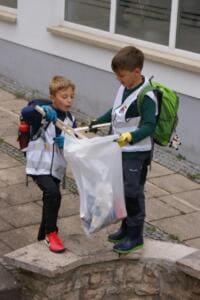 Zwei Jungen in Warnwesten stehen auf einer Mauer und füllen gemeinsam einen durchsichtigen Sack mit Müll.