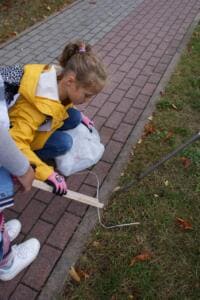 Ein Mädchen in gelber Jacke sammelt kniend mit einer Holzgreifzange Müll am Rand eines gepflasterten Gehwegs.
