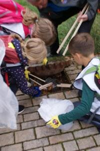 Mehrere Kinder beugen sich mit Holzgreifzangen über einen Gully und holen Müll heraus.