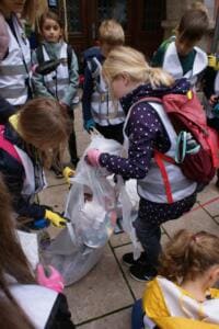 Eine Gruppe Grundschulkinder in Warnwesten steht eng beieinander und füllt Müll in mehrere Beutel.