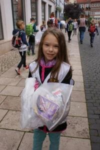 Ein lächelndes Mädchen in Warnweste hält auf einem Altstadt-Gehweg einen vollen durchsichtigen Beutel, dahinter laufen weitere Kinder.