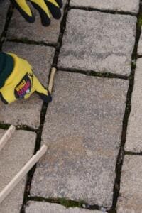 Nahaufnahme von Pflastersteinen, an deren Rand eine Holzgreifzange und Hände in World-Cleanup-Day-Handschuhen nach Müll suchen.