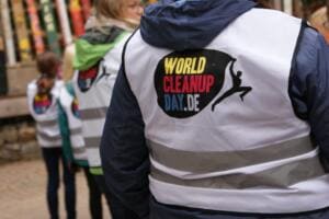 Nahaufnahme des Rückens einer Person in weißer World-Cleanup-Day-Warnweste, im Hintergrund unscharf weitere Kinder in Westen.