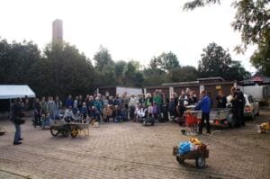 Eine große Gruppe aus Erwachsenen und Kindern steht mit Schubkarren und Anhängern auf einem gepflasterten Platz vor Garagen und einem Pavillon.