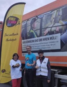 Drei Erwachsene in World-Cleanup-Day-Shirts und Westen zeigen den Daumen hoch vor einem Müllfahrzeug der Stadtwerke Erfurt und einer gelben World-Cleanup-Day-Beachflag.