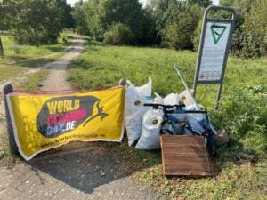 Ein gelbes World-Cleanup-Day-Banner hängt an einem Weg vor vollen weißen Säcken, einer Holzplatte und einem alten Fahrrad.