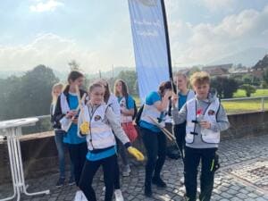 Eine Gruppe Jugendlicher in Warnwesten steht mit Greifzangen auf einer Brücke vor einer Beachflag, im Hintergrund Hügel und Häuser.