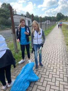 Zwei Mädchen in Warnwesten sammeln mit Greifzange und blauem Sack Müll an einem Gehweg entlang eines Zauns.