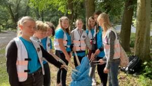 Eine Gruppe Mädchen in Warnwesten steht mit Greifzangen und einem blauen Sack unter Bäumen am Wegrand.