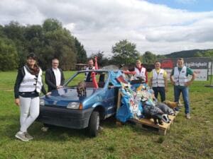 Ein altes blaues Auto ist mit gesammeltem Plastikmüll und Tierfiguren wie Dachs und Fuchs dekoriert, daneben stehen Erwachsene in Warnwesten.