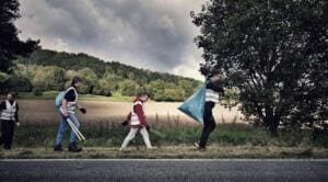 Vier Personen in Warnwesten sammeln mit Greifzangen und Säcken Müll am Rand einer Landstraße vor Feldern und Wald.