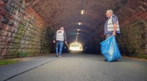 Zwei Männer in Warnwesten sammeln in einem gemauerten Tunnel Müll, einer trägt einen blauen Sack.