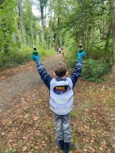 Ein Junge in World-Cleanup-Day-Weste steht von hinten auf einem Waldweg und hält in jeder Hand eine gefundene Glasflasche hoch.