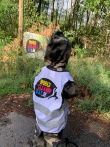 Ein Kind in Tarnkleidung und weißer World-Cleanup-Day-Warnweste steht von hinten an einem Waldweg vor einem Schild mit World-Cleanup-Day-Logo.