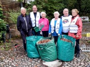 Sechs ältere Erwachsene, einige in World-Cleanup-Day-Westen, stehen auf einem gepflasterten Hof hinter drei grünen Gartensäcken voller Laub und Abfall.