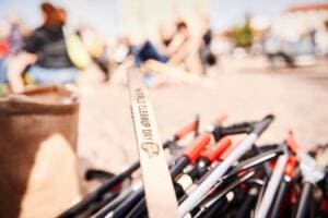 Nahaufnahme eines Bündels Greifzangen und eines Holzstabs mit World-Cleanup-Day-Schriftzug, im unscharfen Hintergrund sitzen Menschen auf einem sandigen Platz.