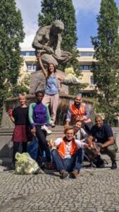 Eine Gruppe junger Erwachsener mit Greifzangen und vollen Säcken posiert an einem Brunnen mit Steinskulptur, eine Frau steht auf dem Brunnenrand und reckt ihre Greifzange.