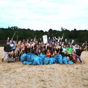 Eine große Gruppe steht jubelnd mit Greifzangen am Sandstrand des Plötzensees hinter einer Reihe voller blauer Müllsäcke.