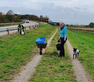 Eine Frau mit Eimer und ein Hund stehen auf einem Feldweg neben einer Schubkarre mit Müllsack, im Hintergrund sammelt ein Mann in Warnweste am Straßenrand.