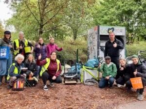 Eine Gruppe Erwachsener zeigt den Daumen hoch, vor ihnen liegen Fundstücke wie ein Fahrradrahmen und Säcke; links eine Tasche mit der Aufschrift Bachpaten Freiburg.