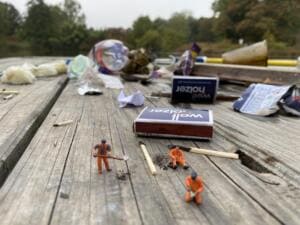 Auf einer verwitterten Holzbank liegen Plastikmüll, Streichholzschachteln und Streichhölzer; davor arbeiten Miniaturfiguren von Bauarbeitern mit Schaufel und Besen.