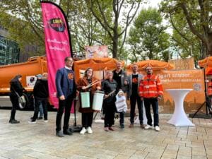 Sechs Erwachsene mit Eimern und Greifzangen stehen vor einem orangefarbenen Müllfahrzeug und einem Banner mit der Aufschrift Stuttgart sauber, daneben eine pinke World-Cleanup-Day-Beachflag.