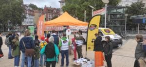 Viele Menschen stehen auf einem Platz vor einem orangefarbenen Pavillon mit Bannern, einer World-Cleanup-Day-Beachflag und einem beklebten Transporter.
