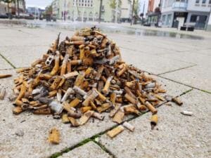 Ein großer Haufen gesammelter Zigarettenstummel liegt auf dem Pflaster eines Platzes, im Hintergrund ein Brunnen und Geschäftshäuser.