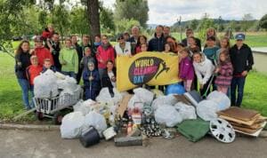 Eine große Gruppe aus Kindern und Erwachsenen steht mit Greifzangen und einer gelben World-Cleanup-Day-Fahne hinter einem großen Haufen Müllsäcke, Flaschen, Pappe und einer Radkappe.