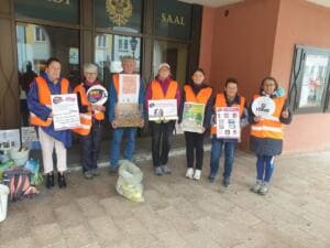 Sieben Erwachsene in orangefarbenen Warnwesten stehen mit Plakaten und World-Cleanup-Day-Schildern vor dem Eingang eines Saals.