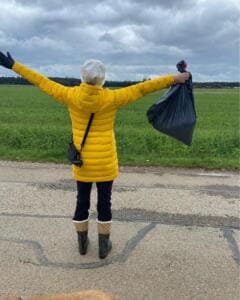 Eine Person in gelber Jacke steht von hinten auf einem Feldweg vor weiten Wiesen und breitet die Arme aus, in einer Hand einen vollen Müllsack.