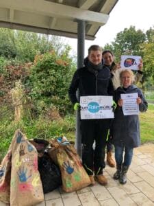 Drei Erwachsene stehen unter einem Unterstand mit Schildern der Aktion unfairmüllt und einem World-Cleanup-Day-Logo, davor liegen mit Handabdrücken bemalte Papiersäcke voller Müll.