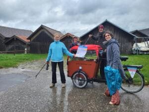 Vier Erwachsene stehen mit Greifzange, Müllsack und einem Plakat neben einem Lastenrad vor alten hölzernen Bootshütten am Starnberger See.