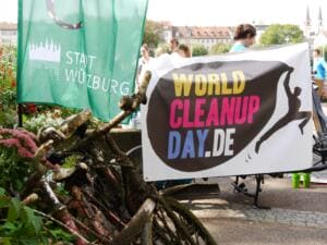 Ein verrostetes, mit Schlamm und Muscheln überzogenes Fahrrad liegt vor einem World-Cleanup-Day-Banner und einer Fahne der Stadt Würzburg.