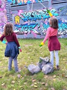 Zwei Mädchen mit Greifzangen stehen von hinten auf einer Wiese vor einer bunten Graffitiwand, neben ihnen gefüllte Müllsäcke.