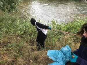 Eine Person in schwarzer Kapuzenjacke beugt sich am steilen Flussufer mit Greifzange ins Gebüsch, vorne hält eine zweite Person einen blauen Müllsack.