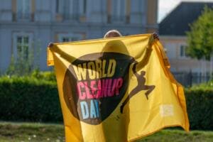 Eine Person hält eine gelbe World-Cleanup-Day-Fahne vor sich ausgebreitet, im Hintergrund Hecken und die Fassade eines Schlosses.
