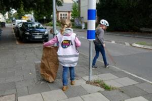 Zwei Mädchen in Warnweste und mit Fahrradhelm sammeln mit Papiersack und Greifzange Müll auf einem Gehweg an einer Straße.