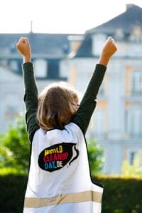 Ein Mädchen in World-Cleanup-Day-Weste reckt von hinten beide Fäuste in die Luft, im unscharfen Hintergrund ein Schloss.