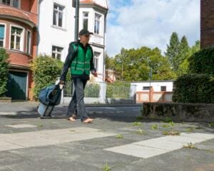 Ein Mann in grüner Warnweste geht barfuß mit Greifzange und einem Rollkoffer voller Müll über einen Gehweg vor Wohnhäusern.