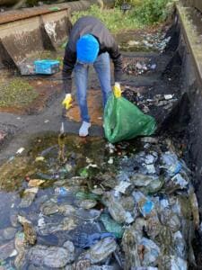 Ein Mann mit blauer Mütze sammelt in einer betonierten, verschmutzten Rinne Plastikflaschen und Müll in einen grünen Sack.