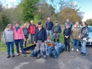 Zwölf Erwachsene stehen am Straßenrand vor Gebüsch neben vollen Müllsäcken und rostigen Fundstücken.