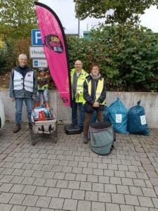 Drei Erwachsene und ein Kind auf einem Bollerwagen stehen neben einer pinken World-Cleanup-Day-Beachflag und vollen blauen Säcken an einer Mauer.