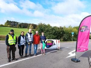 Fünf Erwachsene stehen auf einer Straße vor einem World-Cleanup-Day-Banner an einer Absperrung, rechts eine pinke Beachflag, im Hintergrund Wiesen.
