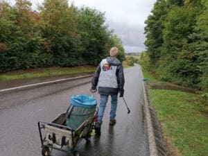 Ein Mann in World-Cleanup-Day-Weste zieht von hinten einen Bollerwagen mit Müllsack eine regennasse Landstraße entlang.
