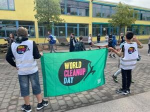 Zwei Schüler in World-Cleanup-Day-Westen halten auf einem Schulhof eine grüne World-Cleanup-Day-Fahne, dahinter weitere Kinder vor einem Schulgebäude.