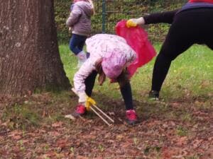 Ein Kind in rosa Jacke sammelt mit einer Holzgreifzange Müll im Laub unter einem Baum, daneben hält eine erwachsene Person einen pinken Sack.